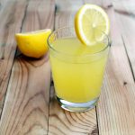 Лимонный сок в стакане, на который надета долька лимона, стоит на деревянном столе перед половинкой плода лимона